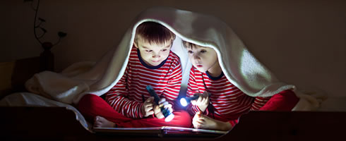 Photo of two boys in pyjamas reading by torchlight uner a blanket